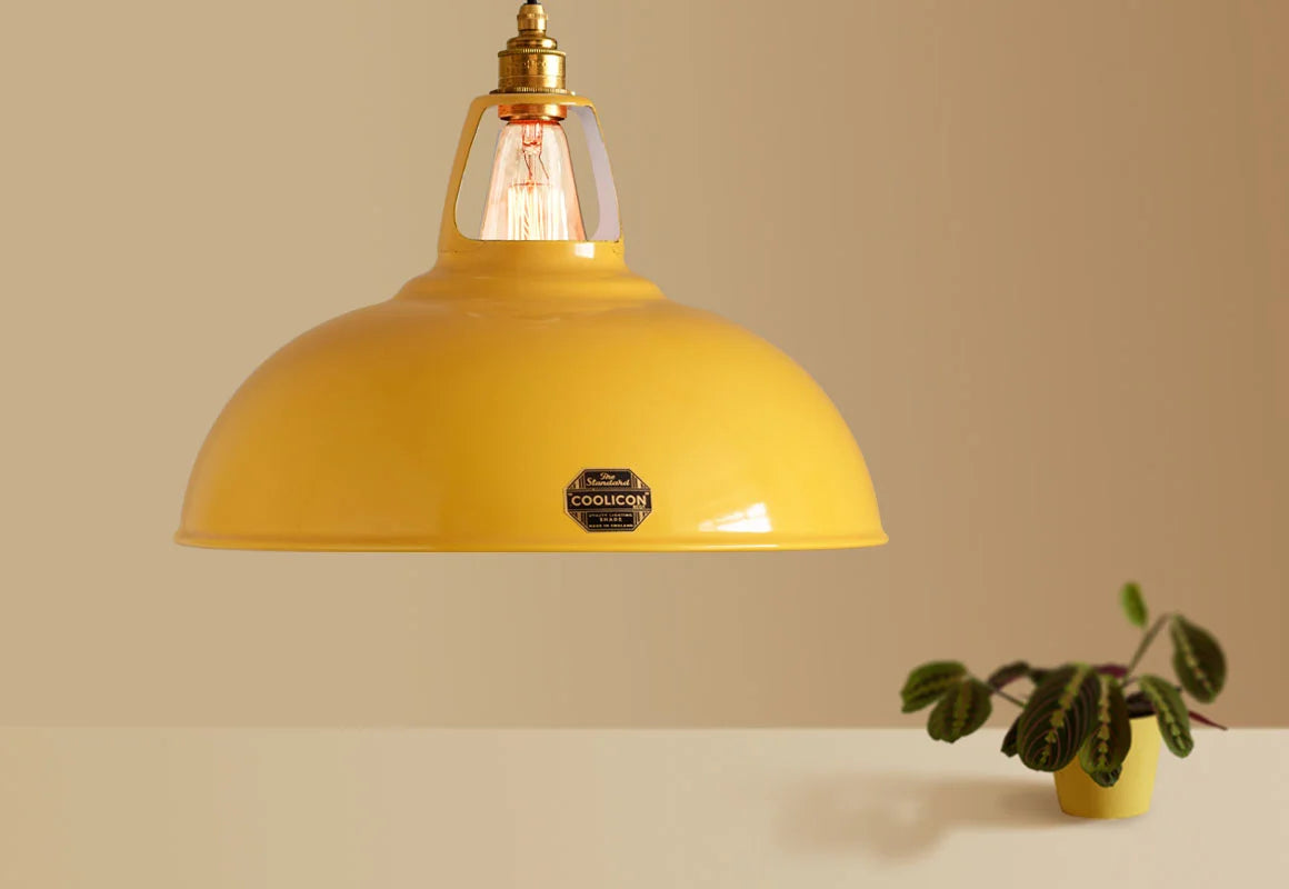 A Large Coolicon Deep Yellow lampshade hanging over a plinth. Below the shade is a pinstripe calathea plant in a yellow plant pot and a pile of yellow books and notebooks with a yellow cup on top